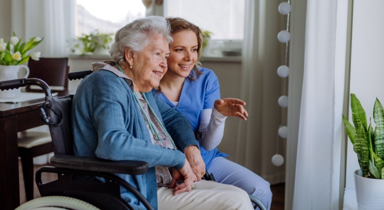 Eine junge Pflegekraft in blauer Kleidung sitzt neben einer Seniorin im Rollstuhl und blickt mit ihr l&auml;chelnd aus dem Fenster. Die Atmosph&auml;re ist herzlich und zugewandt.